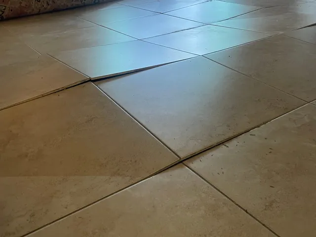 Buckled floor tiles tenting upward along a grout line in a living room